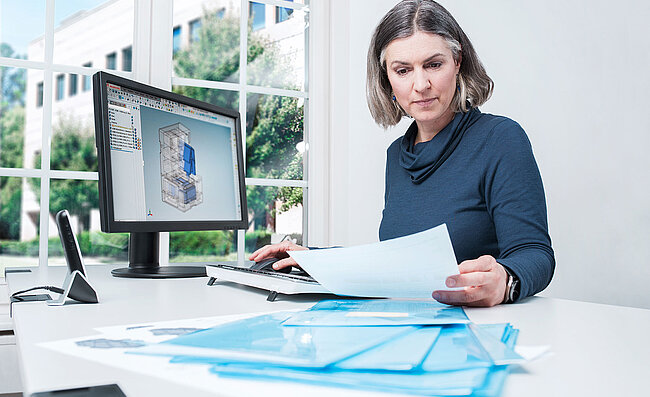 Žena sedí u stolu před počítačem, na obrazovce jsou technické výkresy, před sebou má dokumenty