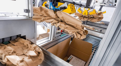 
				Automated packing system with brown paper being dispensed into a cardboard box for cushioning and protection during shipping.